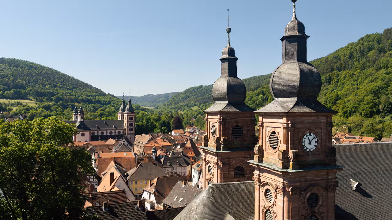 drohnenaufnahme zweier kirchtürme mit turmuhr und kloster im hintergrund in amorbach bei sonnenschein