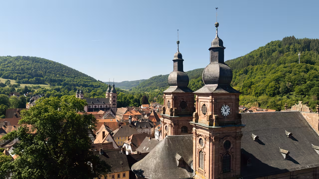 drohnenaufnahme zweier kirchtürme mit turmuhr und kloster im hintergrund in amorbach bei sonnenschein