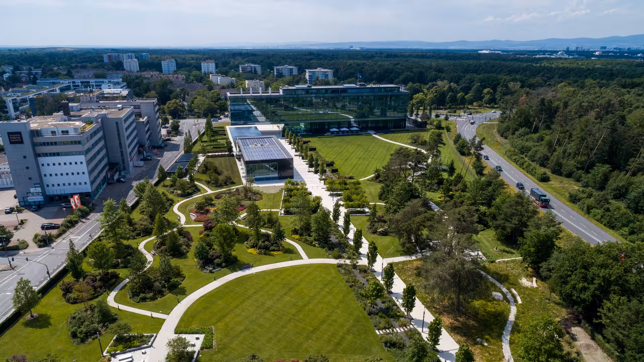 drohnenaufnahme eines modernen parks mit bäumen, rasenflächen und sitzgelegenheiten sowie einem modernen bürogebäude mit glasfassade im hintergrund in dreieich