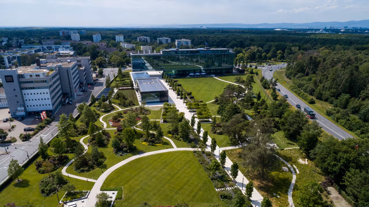 drohnenaufnahme eines modernen parks mit bäumen, rasenflächen und sitzgelegenheiten sowie einem modernen bürogebäude mit glasfassade im hintergrund in dreieich